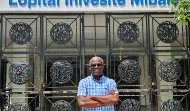 Dr. Lambert is wearing a shortsleeved blue collared shirt, standing with his arms crossed in front of the gate to one of Zanmi Lasante's clinics