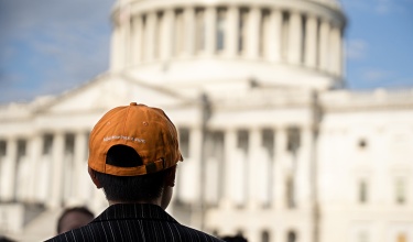 Person in an orange hat faces away from the camera with the U.S. Capitol building blurry in the background. The back of the hat says "Injustice has a cure."