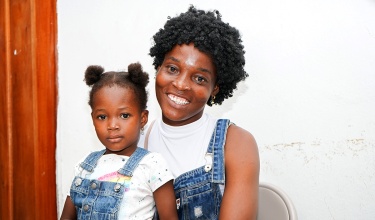 Tatiana sits on her mother Thérèse's lap, both are wearing denim overalls with white shirts underneath