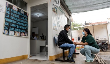 A person in a green shirt wearing a mask squats on the ground looking up at the person in front of them in a black jacket also wearing a mask sitting on a bench outside a clinical room, gripping hands