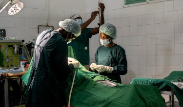 Doctors and nurses in green scrubs stand around a hospital bed as they perform a C-section on a patient.