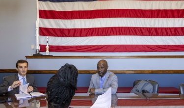 Three people talk with one another while sitting at tables underneath an American flag.