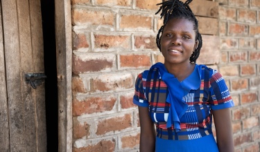 Bernadette Daluni, a 20-year-old woman, stands in front of a brick house wearing a bright blue patterned shirt.