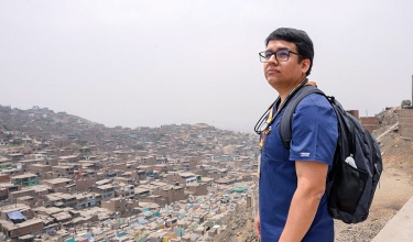 Dr. Santiago Palomino, wearing navy scrubs, a black backpack, black rimmed glasses, and a stethoscope around his neck, looks over a town from above