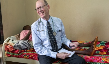 A man in glasses wearing a blue shirt, dark tie, and dark pants with a stethoscope around his neck sits on the edge of a hospital bed with colorful sheets and smiles