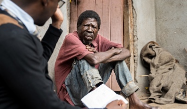 Saulos Metio, a man in his 60s, sits on the ground in a red shirt and blue jeans. He looks at another man, Charles Marshal, who is in the foreground of the photo.