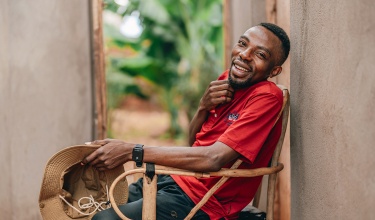 A man in a bright red shirt sits in a wooden chair, smiling. His right hand is brushing his chin, and his left hand is clutching a hat.