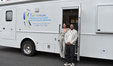 A large, white RV takes up the majority of the screen. In the RV's doorway stands a woman and a man. The woman has her arm propped on the man's shoulder.