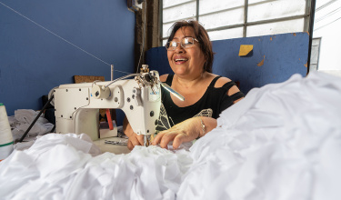 María smiles while sitting at a sewing maching working with billowy white fabric