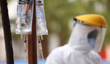 Image shows a clear medical bag hooked up to drip. In the background, there is a blurred health care worker wearing white protective equipment and a mask.
