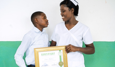 A woman in a nurse uniform smiles at her son while holding her nursing diploma.