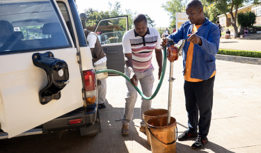 Two men stand to the right of a white van, filling it with fuel out of yellow buckets.