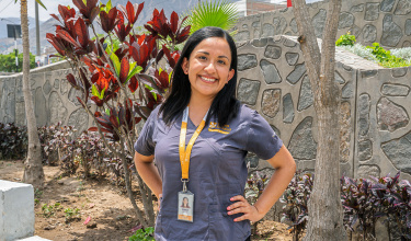 Daniela Puma Abarca smiles, facing the camera, wearing a grey polo and an orange lanyard in front of a stone wall