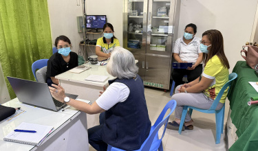 A group of health care workers sit around a table wearing masks. A person with a navy vest gestures toward a computer screen.