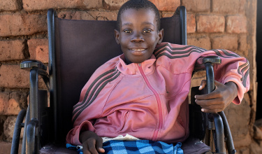 A young girl in a pink jacket smiles as she sits in a wheelchair. 