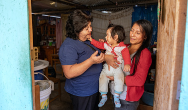 a woman in a red cardigan to the right holds a child in a white and red onesie who is reaching out to touch the cheek of a woman in a navy blue polo