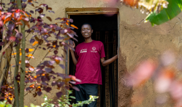 A young man stands in a doorway in a red shirt and smiles.