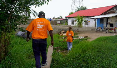 Two people walk along a path through grass to a home up ahead while wearing bright orange shirts that say "health is a human right"